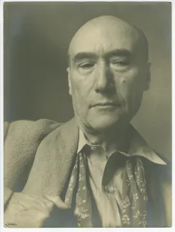 portrait photographique en buste d’André Gide, avec foulard fleuri, de face, regardant vers le bas, main droite sur le col, en contre-plongée, au Vaneau