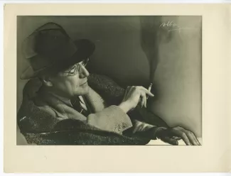 portrait photographique en buste d’André Gide, avec chapeau et lunettes, assis, de profil à droite, une cigarette dans la main droite, la gauche posée sur une table, mars 1939