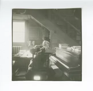 portrait photographique d’André Gide, avec lunettes et bonnet, à califourchon sur une chaise à côté de son piano, fumant, au Vaneau