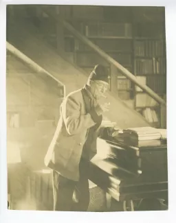 portrait photographique d’André Gide, avec lunettes et bonnet, debout devant son piano, s'allumant une cigarette, au Vaneau