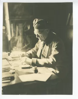portrait photographique d’André Gide, avec lunettes et bonnet, assis à son bureau, écrivant et fumant, au Vaneau