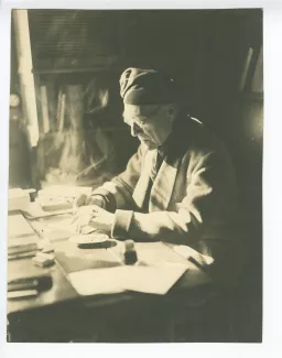 portrait photographique d’André Gide, avec lunettes et bonnet, assis à son bureau, écrivant et fumant, au Vaneau