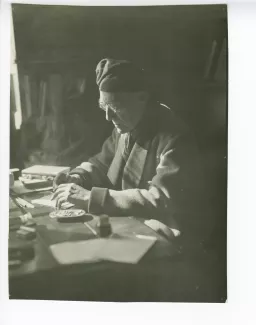 portrait photographique d’André Gide, avec lunettes et bonnet, assis à son bureau, écrivant et fumant, au Vaneau