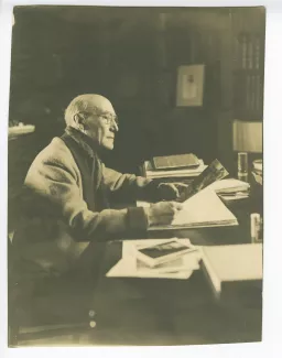 portrait photographique d’André Gide, avec lunettes, assis à son bureau, regardant une grande image, au Vaneau