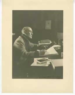 portrait photographique d’André Gide, avec lunettes, assis à son bureau, regardant une grande image, au Vaneau