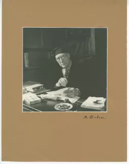 portrait photographique d’André Gide, avec lunettes et bonnet, assis à son bureau, une lettre à la main, fumant, au Vaneau