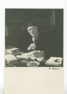 portrait photographique d’André Gide, avec lunettes et bonnet, assis à son bureau, une lettre à la main, fumant, au Vaneau