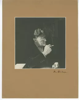 portrait photographique d’André Gide, avec lunettes et bonnet, de ¾ à droite, assis à son bureau, fumant, au Vaneau