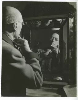 portrait photographique en buste d’André Gide, avec lunettes, de dos, face à un miroir, faisant son nœud de cravate, chez Richard Heyd, hiver 1947