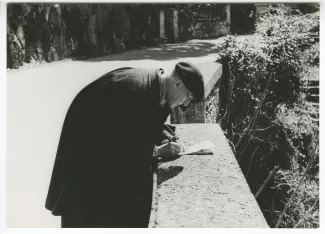 photographie d’André Gide, avec lunettes et béret, écrivant penché sur un muret, en extérieur, avril 1947