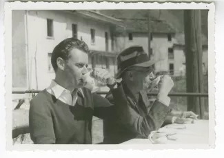 photographie d’André Gide, avec lunettes et chapeau, et Jean Lambert, attablés, buvant tous deux dans une tasse, en extérieur, avril 1947