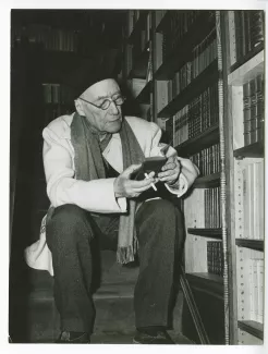 portrait photographique d’André Gide, avec lunettes et bonnet, assis sur un escalier au pied de sa bibliothèque, lisant, au Vaneau, février 1950 ; reportage pour Paris-Match