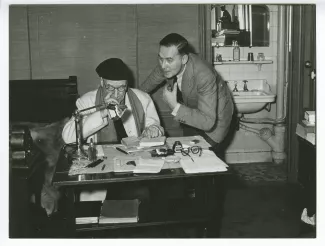 photographie d’André Gide, avec lunettes et bonnet, assis à un bureau, et Pierre Herbart, au Vaneau, février 1950 ; reportage pour Paris-Match