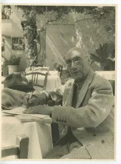 photographie d’André Gide, écrivant, assis à une table, en extérieur, avril-mai 1950