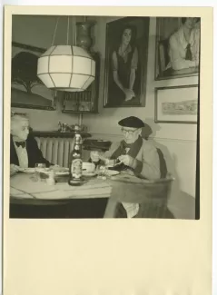 photographie d’André Gide, avec lunettes et béret, et Maria Van Rysselberghe, dans l'appartement de Maria, simulant un déjeuner, lors du tournage du film Avec André Gide de Marc Allégret, janvier 1951