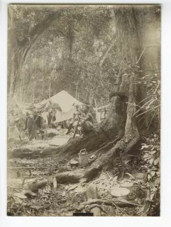 photographie de quatre autochtones préparant la cuisine devant un campement ; dans le fond, on distingue à droite le capitaine Binger et à gauche le lieutenant Braulot, 29-30 janvier 1892